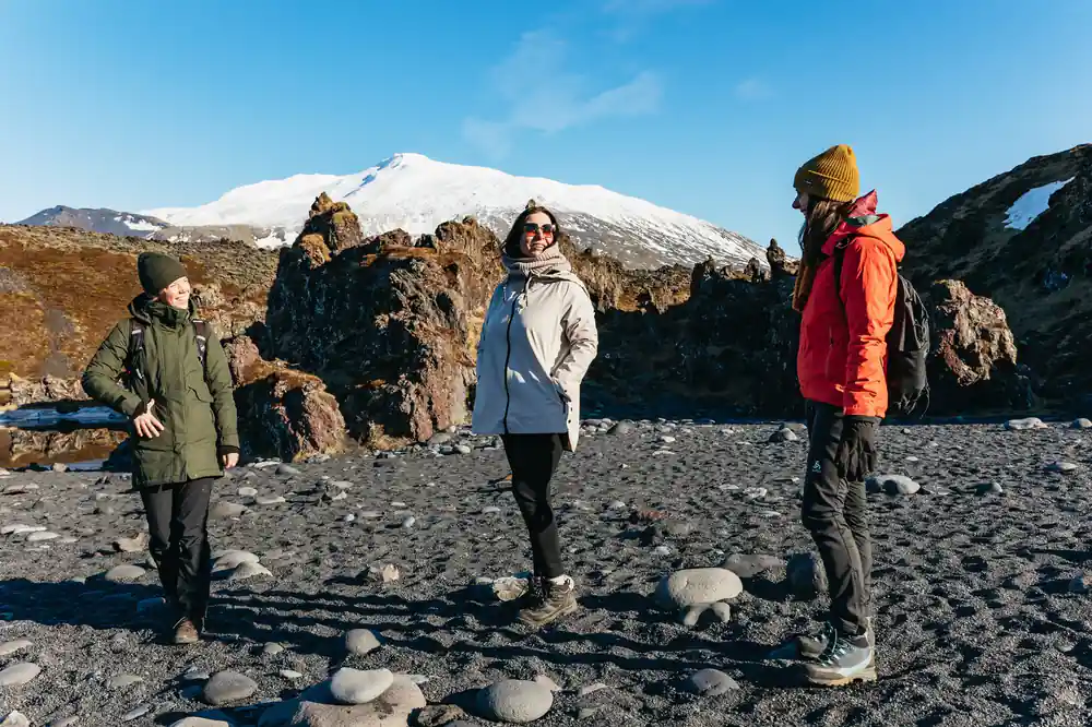 People standing on rocky black sand beach with snow mountain