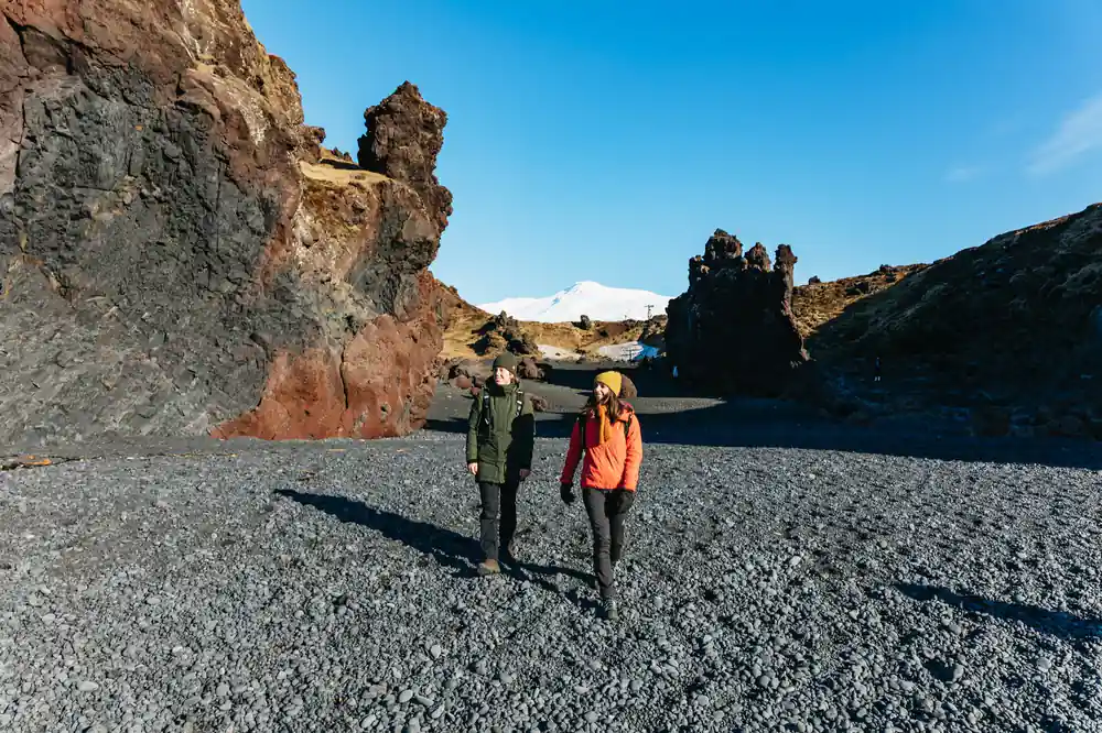 Two people walking on gravel beside tall rock pillar